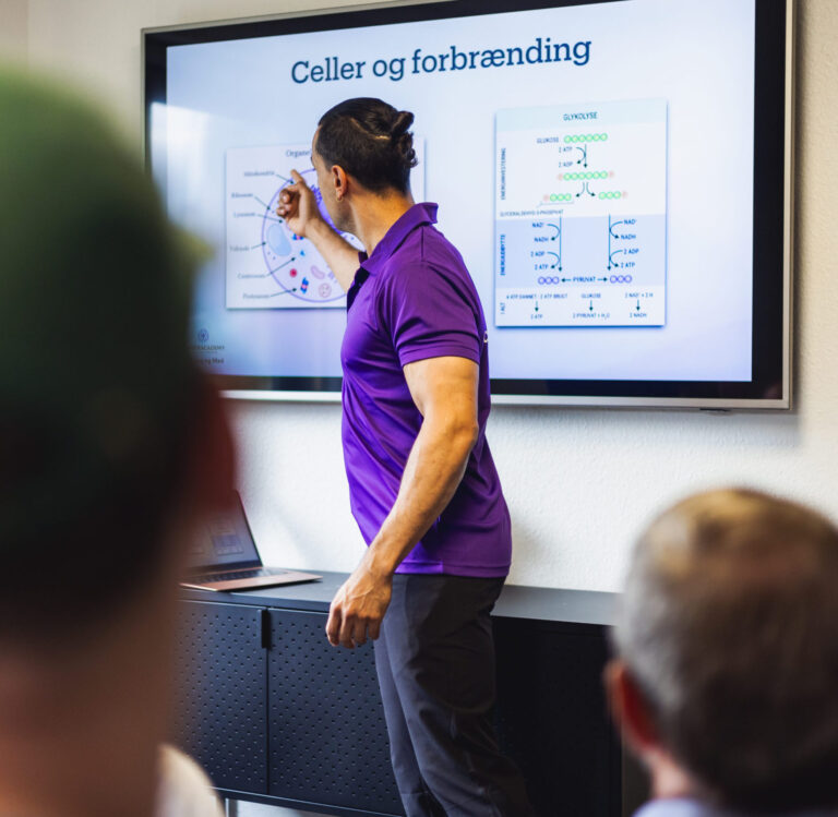 Rune underviser i celleforbrænding foran en skærm med en præsentation om celler og forbrænding. To tilhørere ses i forgrunden, mens personen peger på diagrammer, der illustrerer biokemiske processer.