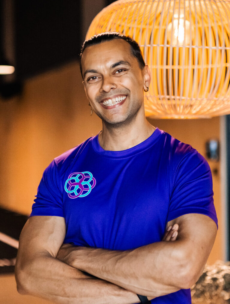 Rune iført en lilla t-shirt med logo står med armene over kors i et fitnesscenter. I baggrunden ses en stor, dekorativ lampe, der skaber en hyggelig og moderne atmosfære.