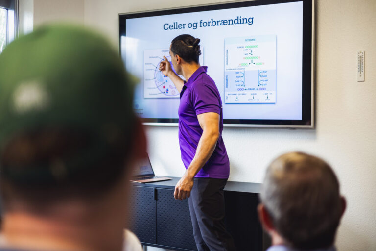 Rune underviser i celleforbrænding foran en skærm med en præsentation om celler og forbrænding. To tilhørere ses i forgrunden, mens personen peger på diagrammer, der illustrerer biokemiske processer.